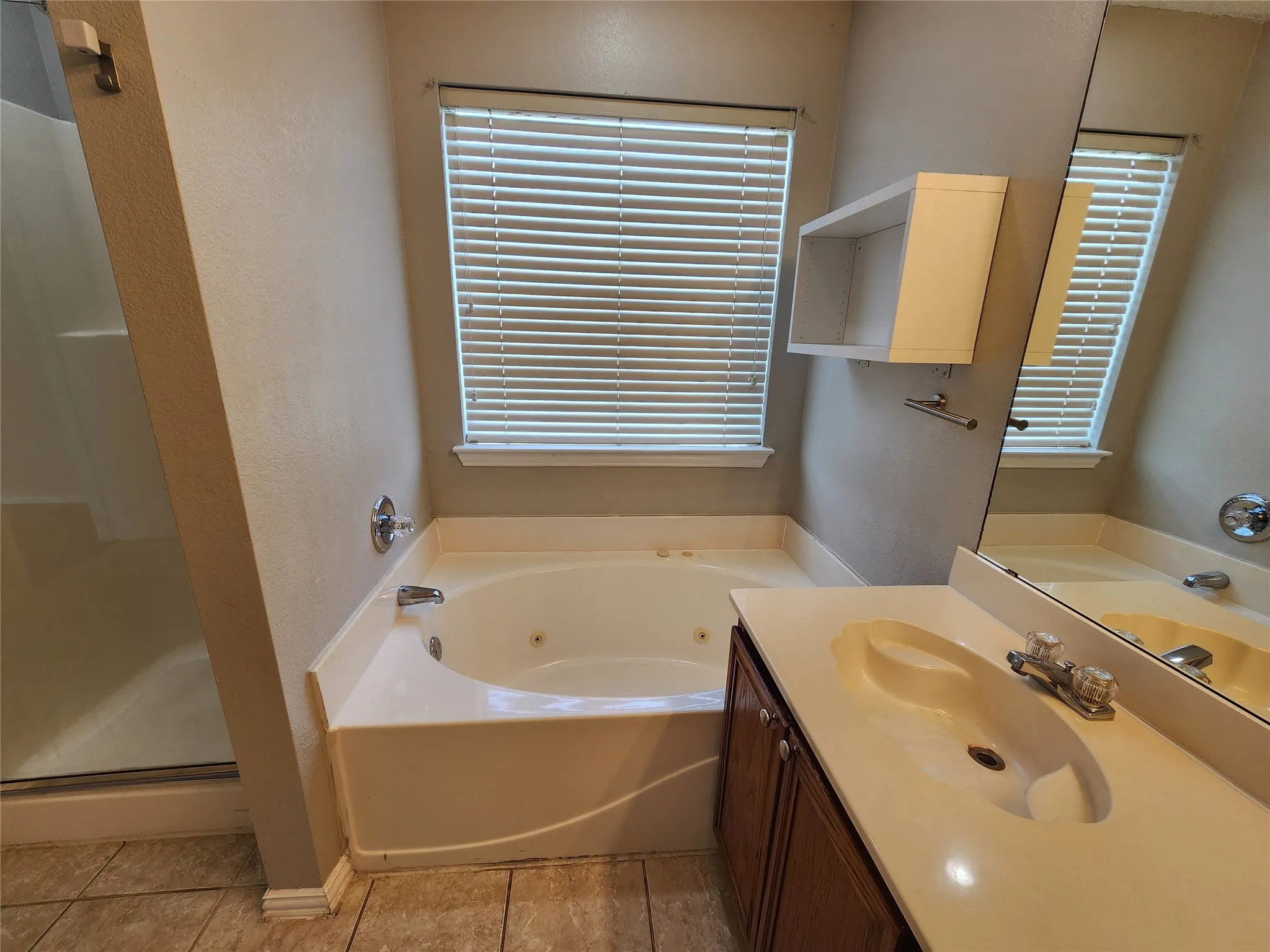 Bathroom featuring a built-in jetted tub, a single vanity with an integrated sink and wood-finish cabinetry, a window with horizontal blinds, a mounted mirror, and tile flooring