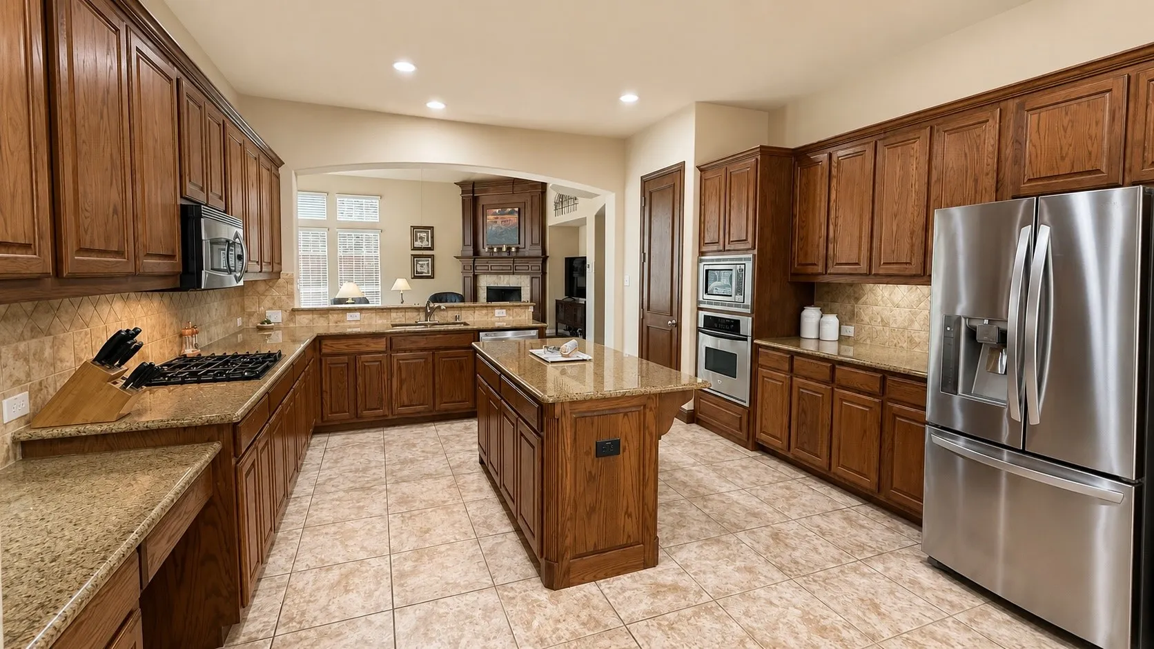 Spacious kitchen featuring a central island, wood cabinetry, granite countertops, tile flooring, and stainless steel appliances including a built-in oven and microwave