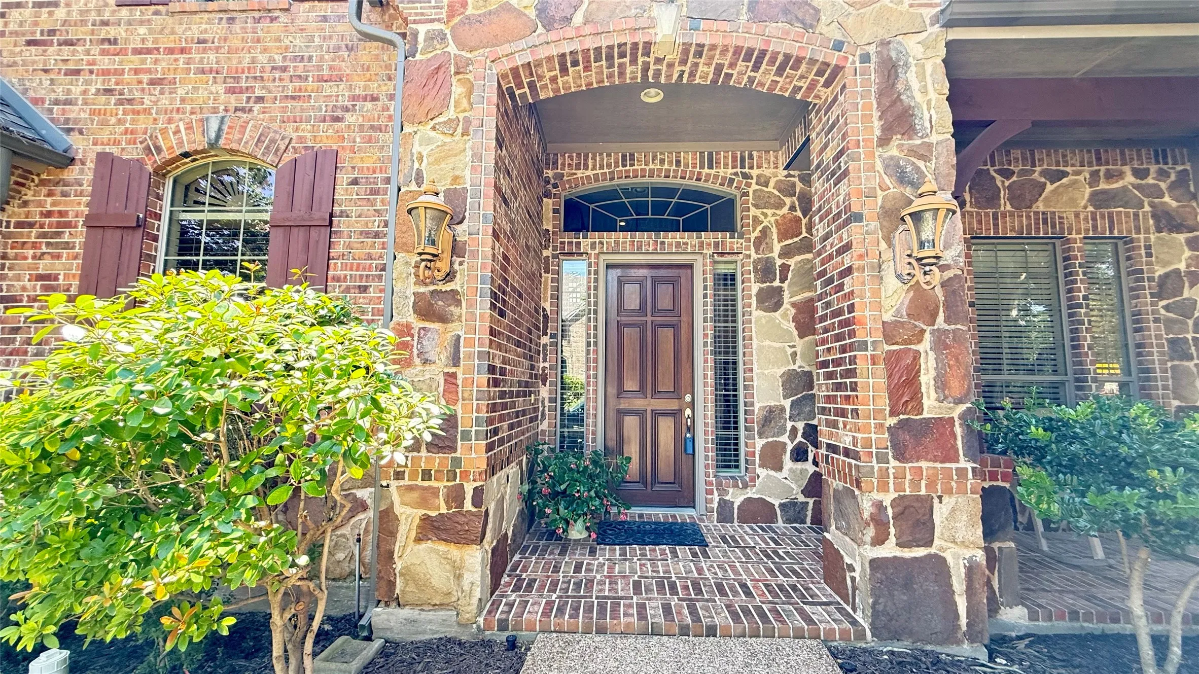Architectural entry featuring a wood paneled door, stone and brick facade, brick archway, decorative outdoor sconces, and arched transom window