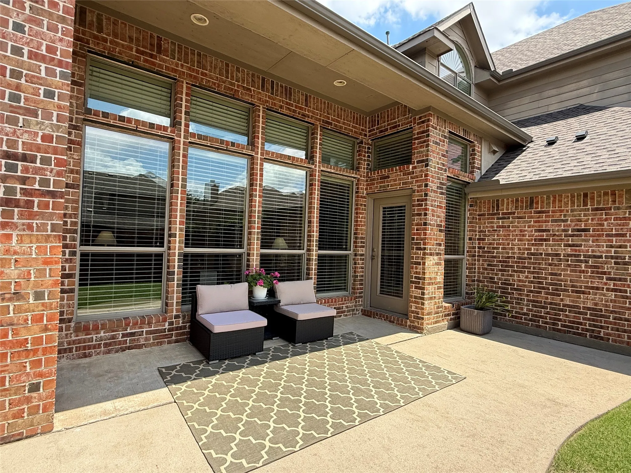 Expansive brick exterior featuring a covered patio, multiple large windows with blinds, a glass-paneled door, and a shingled roofline