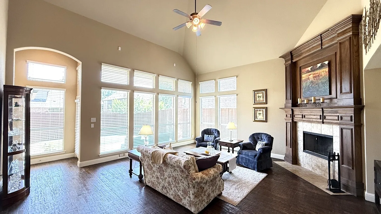 Spacious living area featuring a vaulted ceiling with a ceiling fan, a prominent wood-finished fireplace with tile surround, wood-finish flooring, and a wall of windows with blinds