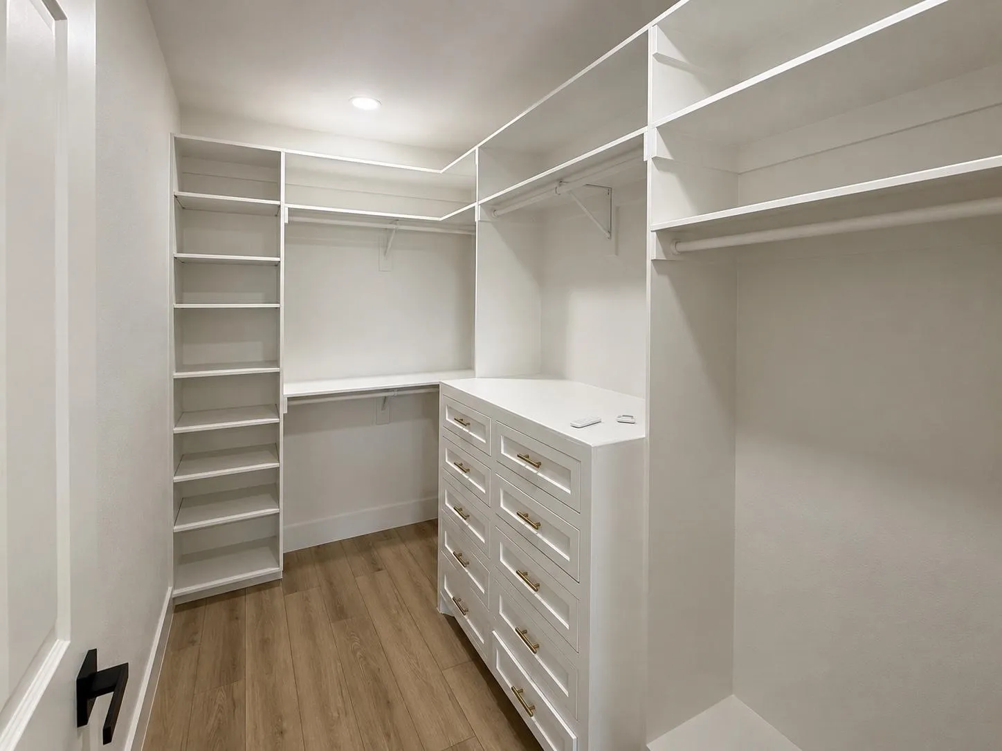 Walk-in closet featuring built-in shelving, multiple hanging rods, a central dresser island with multiple drawers and gold-finish hardware, and wood-finish flooring
