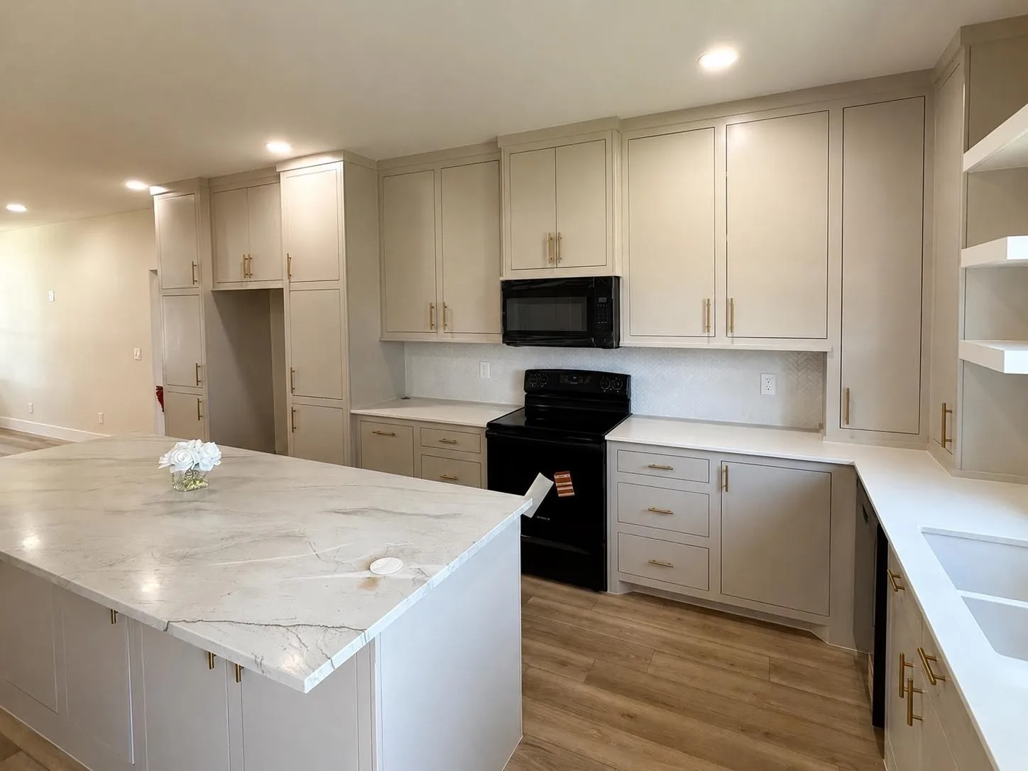 Spacious kitchen featuring a large island with a light-toned countertop, extensive light-colored cabinetry with gold-tone hardware, wood-finish flooring, a black oven range, and a white tile backsplash