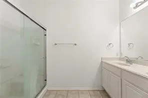 Shower with clear glass enclosure, white walls, double vanity with integrated sinks, large mirror, and wood-finish flooring