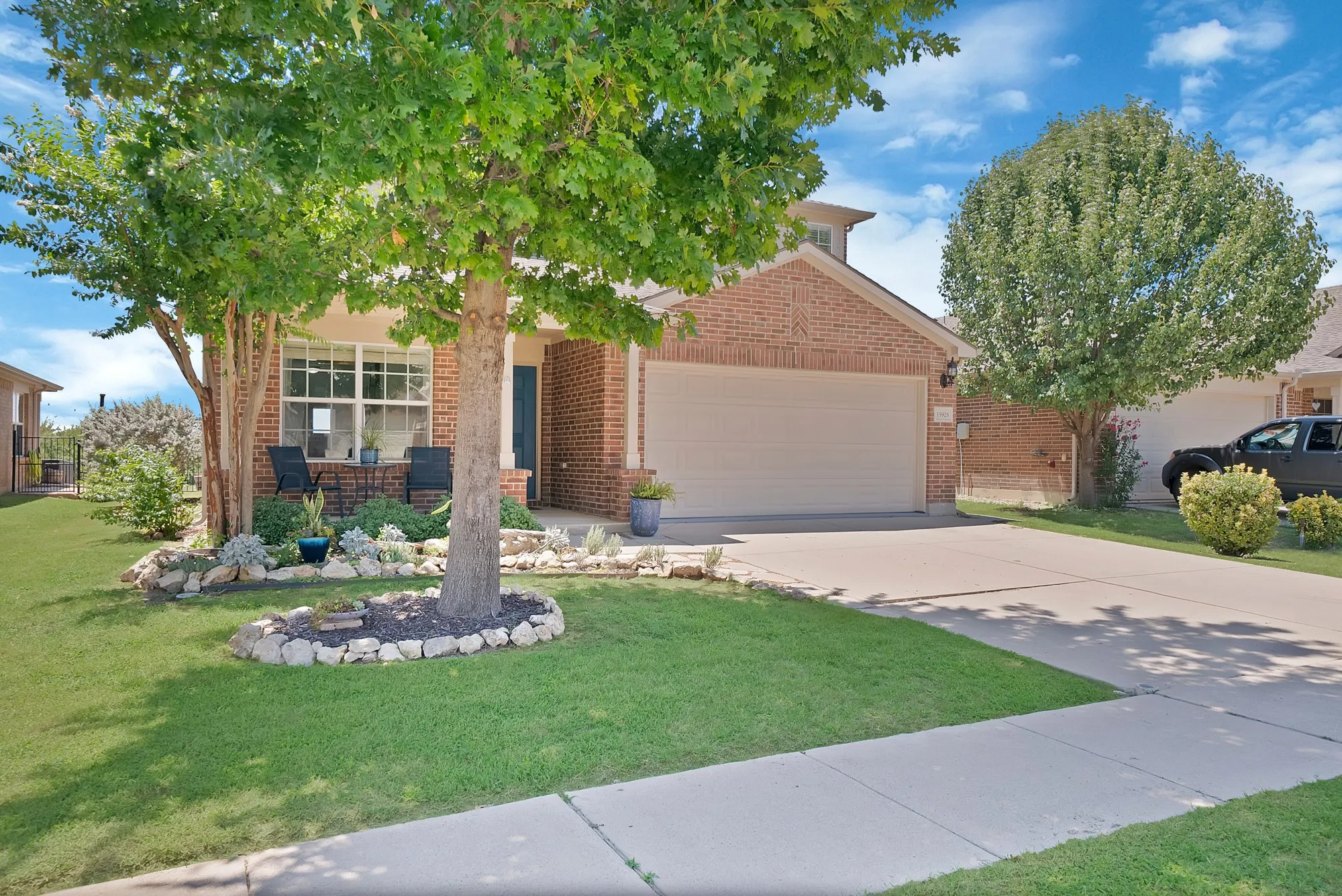 Brick facade home featuring a prominent two-car garage, manicured lawn, established front landscaping with stone accents, mature trees, and a concrete driveway