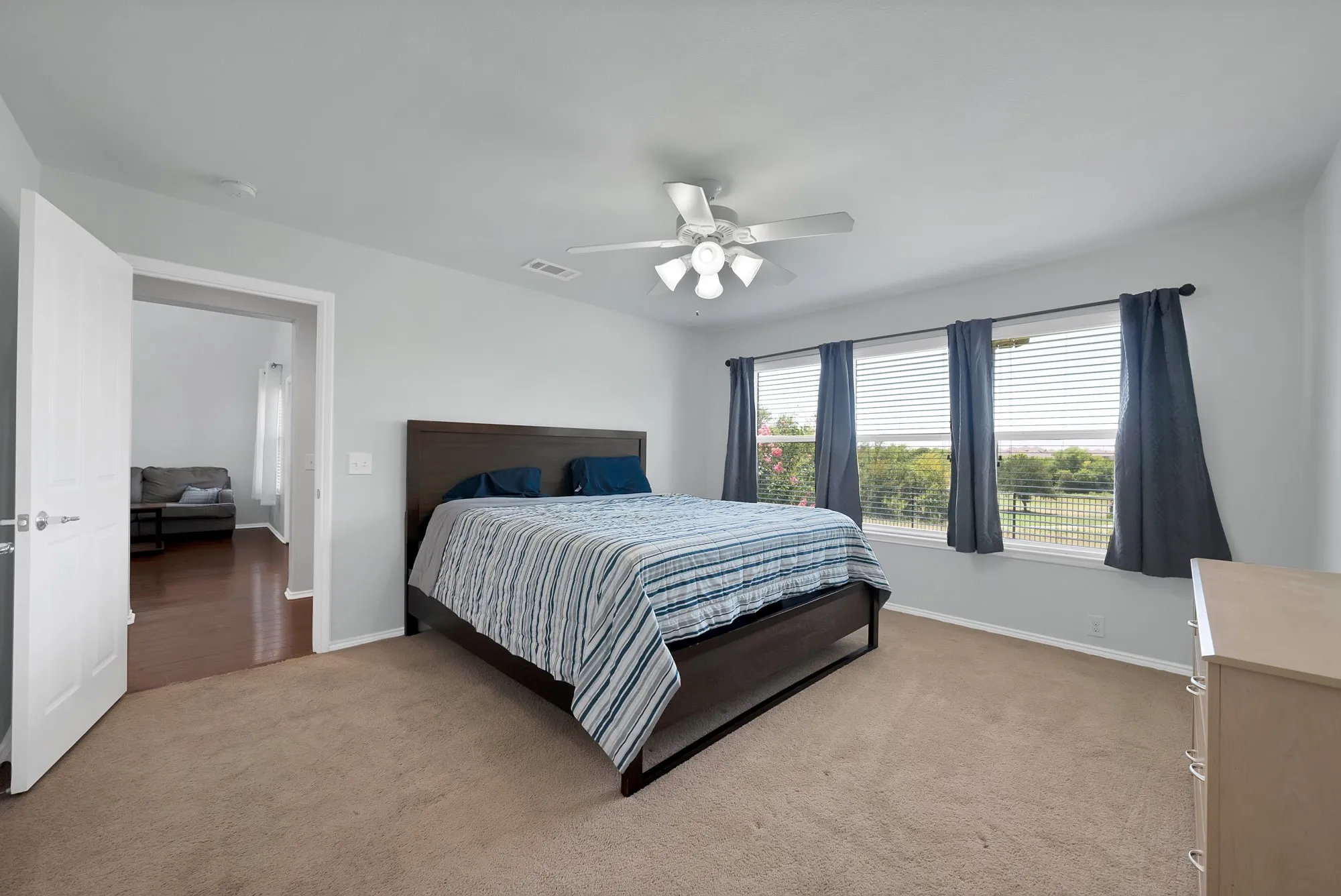Spacious bedroom featuring neutral carpeting, a ceiling fan with integrated lighting, and a series of three windows equipped with blinds