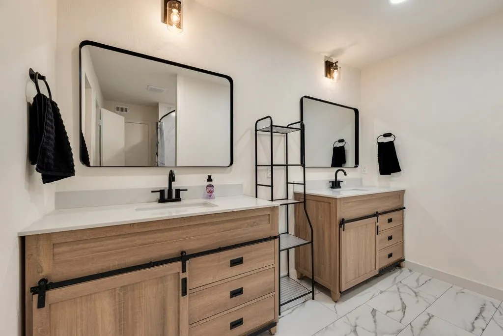 Dual vanity bathroom featuring light wood-finish cabinetry with barn door style sliding panels, white countertops, and rectangular under-mount sinks