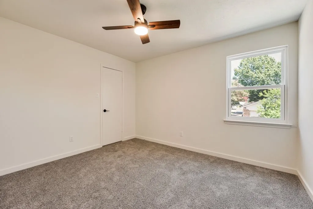 Neutral-toned room featuring a single window with exterior views, light gray carpet flooring, a white paneled door with a black knob, and a ceiling fan with a light fixture
