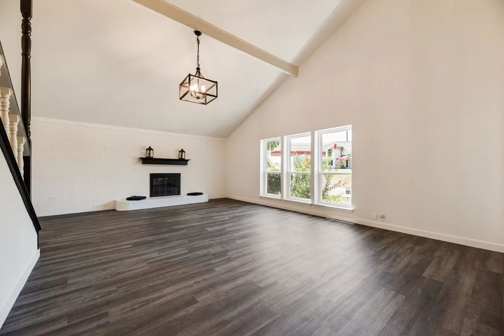 Spacious living area featuring a vaulted ceiling with exposed beam, wood-finish flooring, a white-painted brick fireplace, and a trio of windows