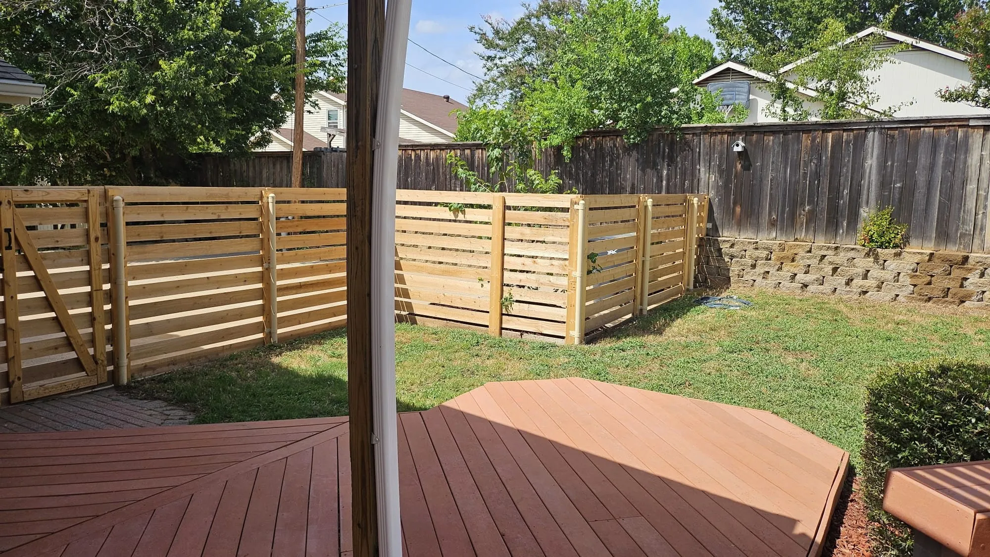 Spacious outdoor area featuring a multi-level composite deck, horizontal slat wood fencing, a grass lawn, and a block retaining wall