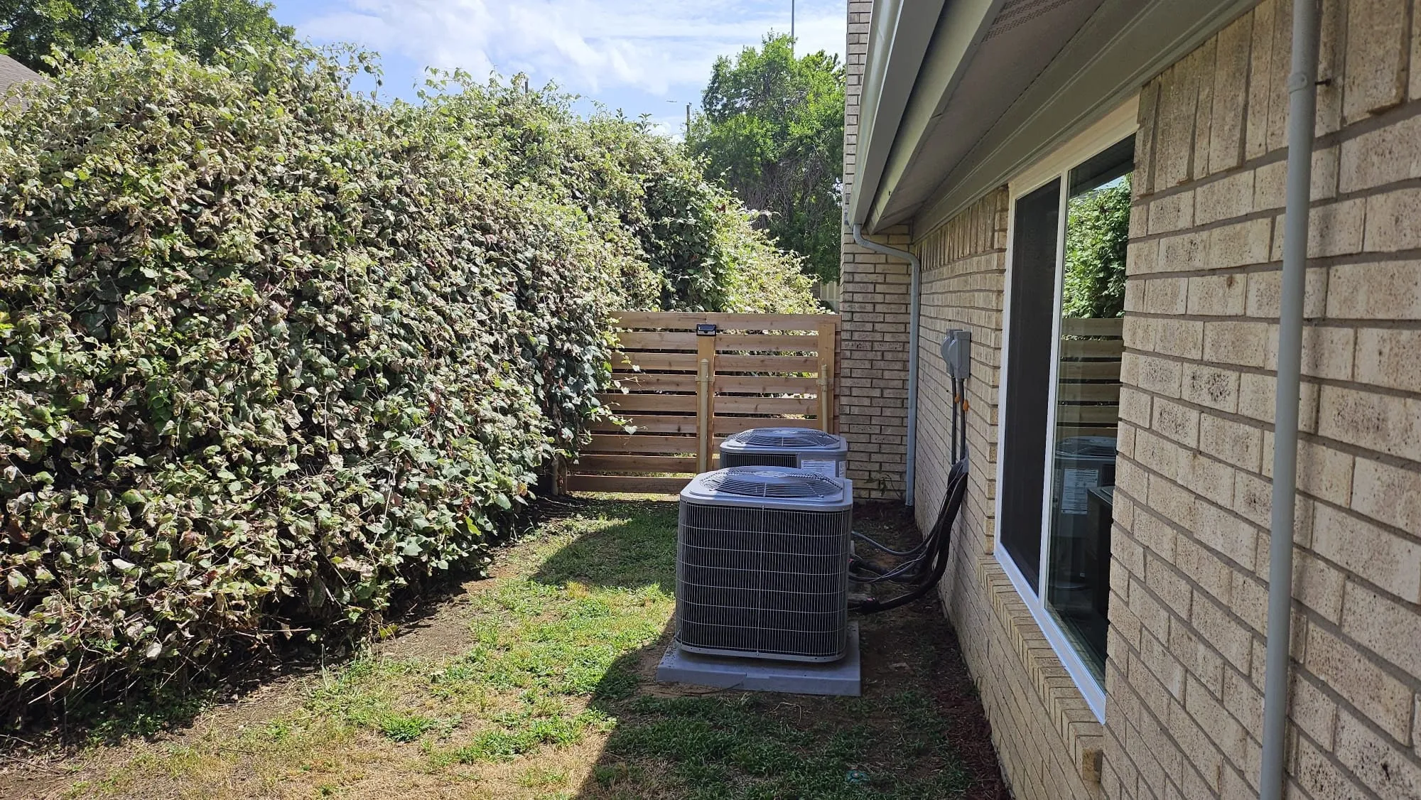 Side yard featuring a mature green hedge, horizontal slat wood fence, two outdoor HVAC units on concrete pads, and a brick exterior with a white-framed window