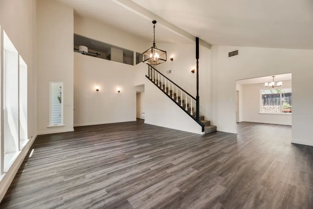 Spacious open-concept living area featuring vaulted ceilings, wood-finish flooring, a contemporary chandelier, a staircase with white spindles, and ample natural light from multiple windows