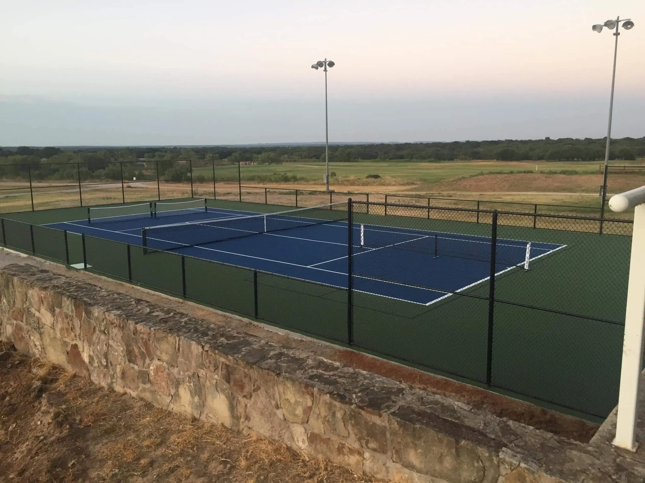 Multi-court tennis facility featuring blue playing surfaces, green surrounds, and perimeter fencing
