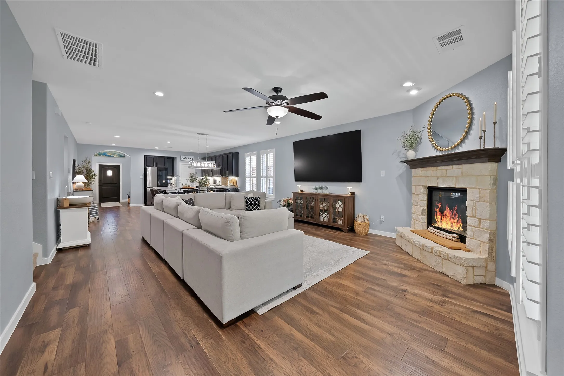 Spacious open-concept living area featuring wood-finish flooring, a corner stone-faced fireplace with wood mantel, recessed lighting, and a ceiling fan