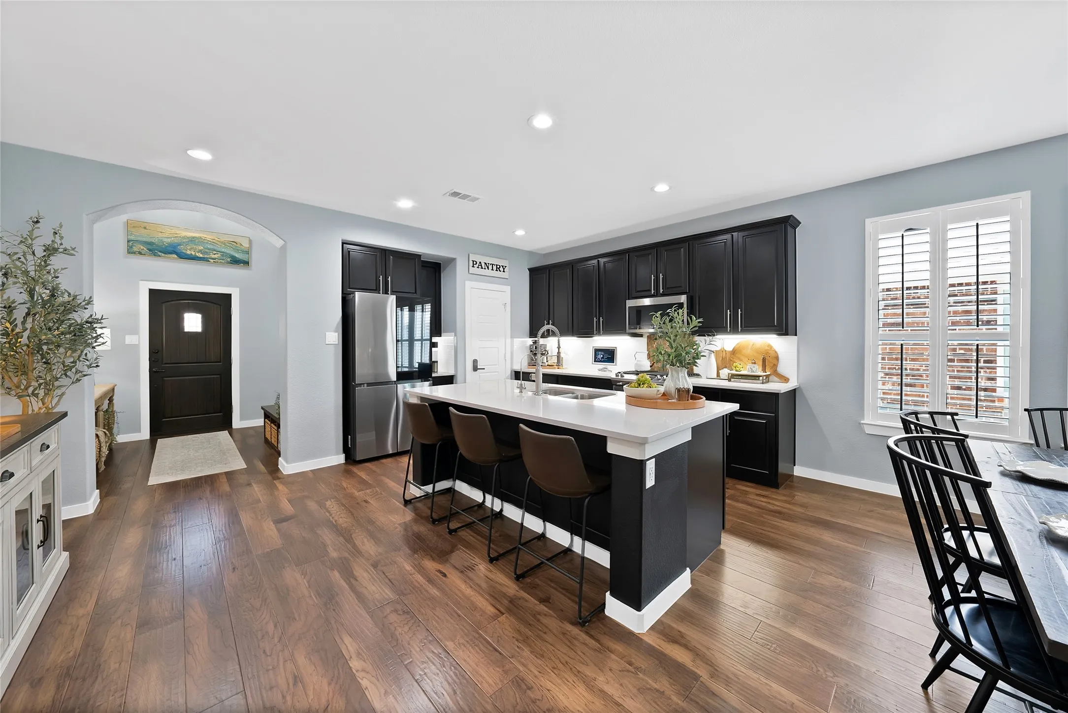 Open-concept kitchen with dark wood-finish flooring, a large island featuring bar seating and a light-toned countertop, black cabinetry, and stainless steel appliances