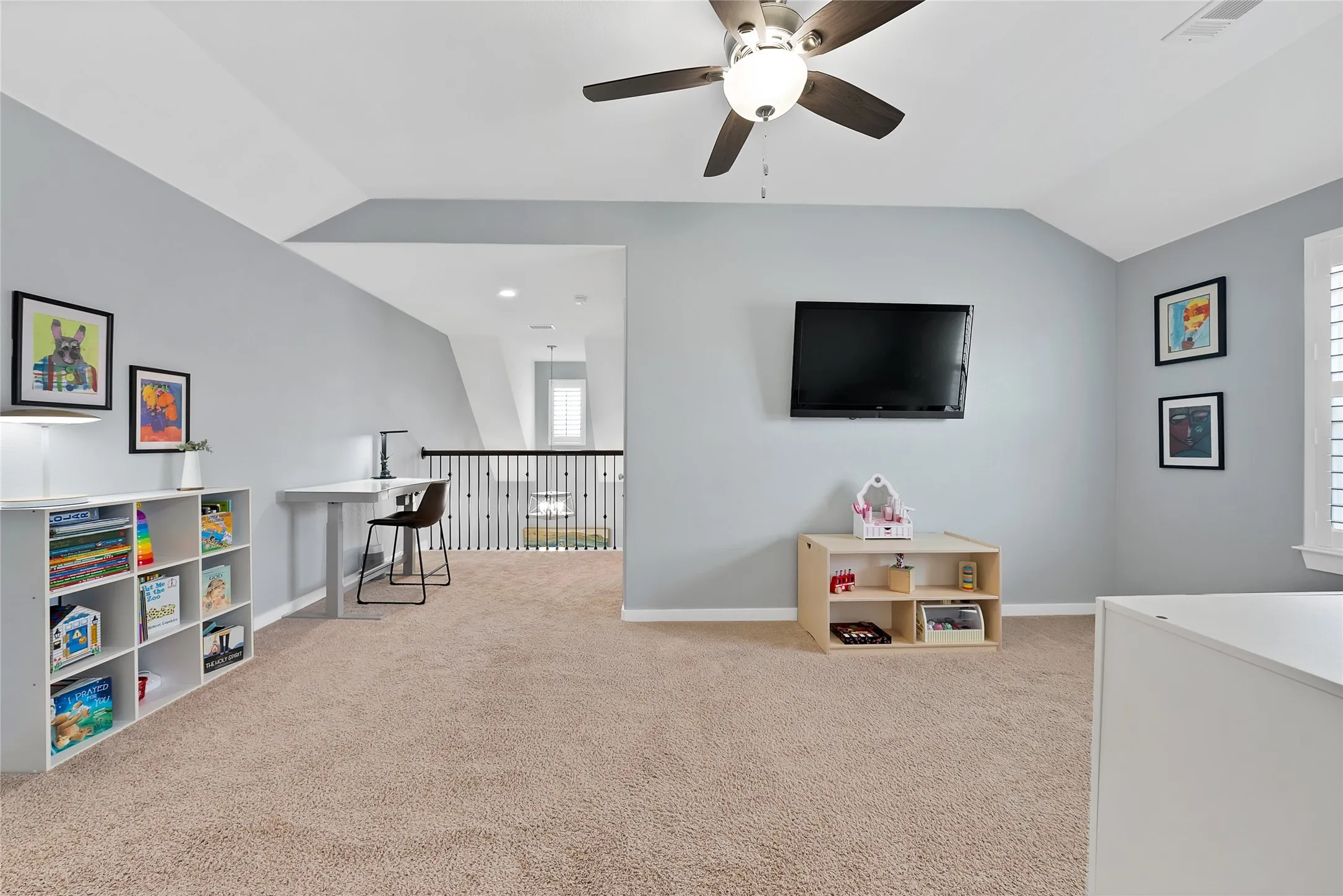 Carpeted loft area featuring a vaulted ceiling with a ceiling fan, light gray walls, an open railing overlooking a lower level, and a window with plantation shutters