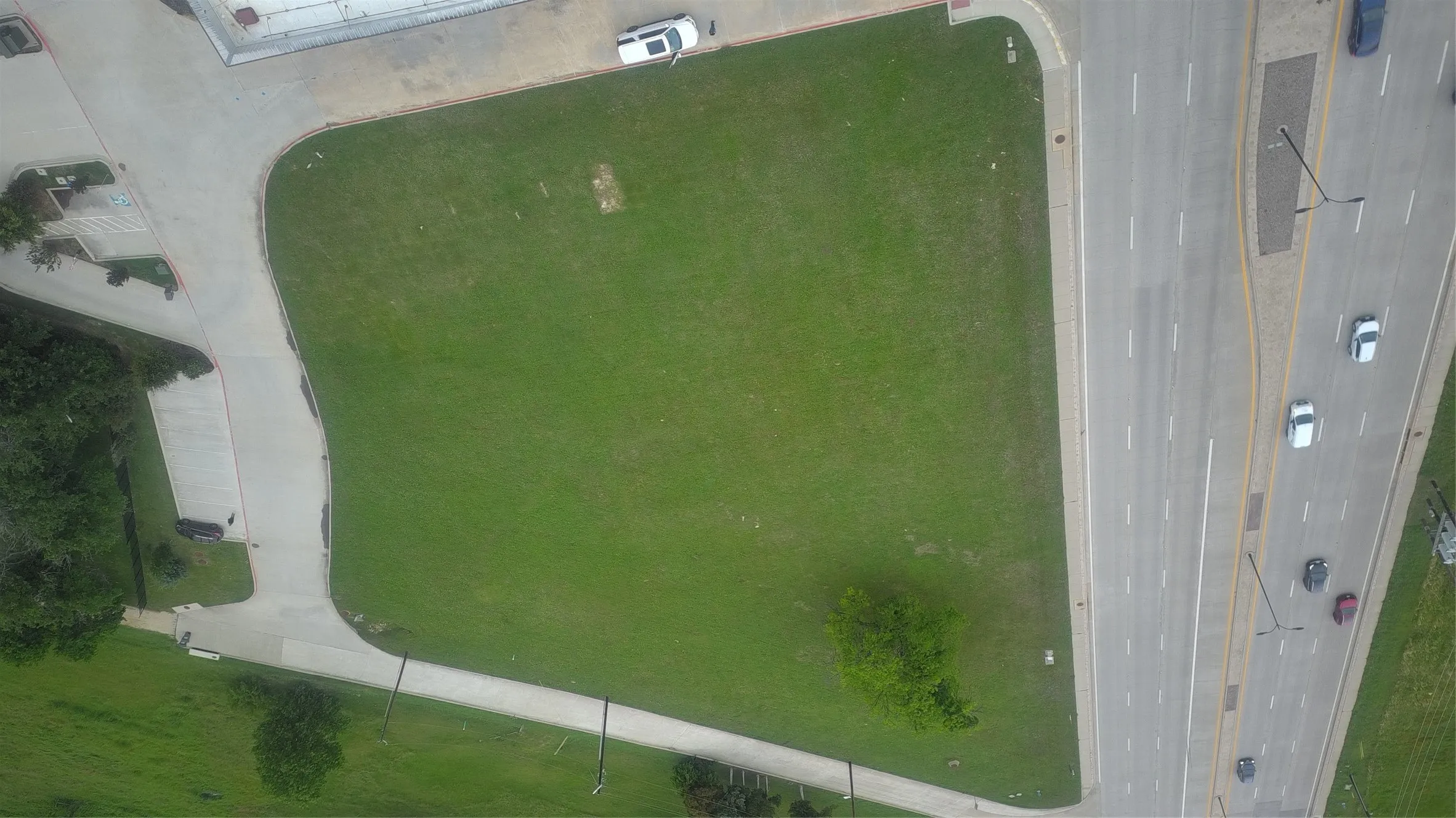 Expansive grass lot with mature trees, bordered by a multi-lane paved road