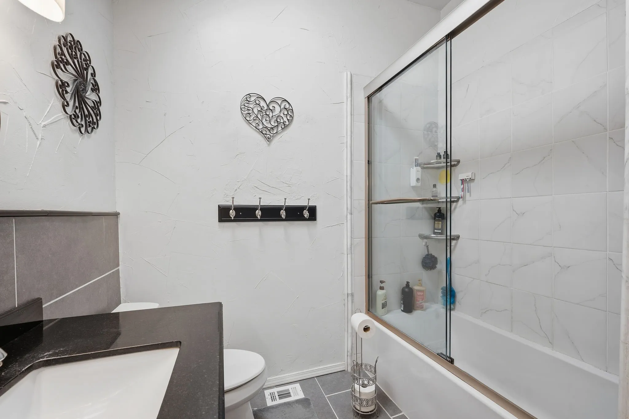Bathroom featuring a white subway tile tub surround, clear glass shower enclosure, dark countertop with an integrated sink, and grey floor tiling