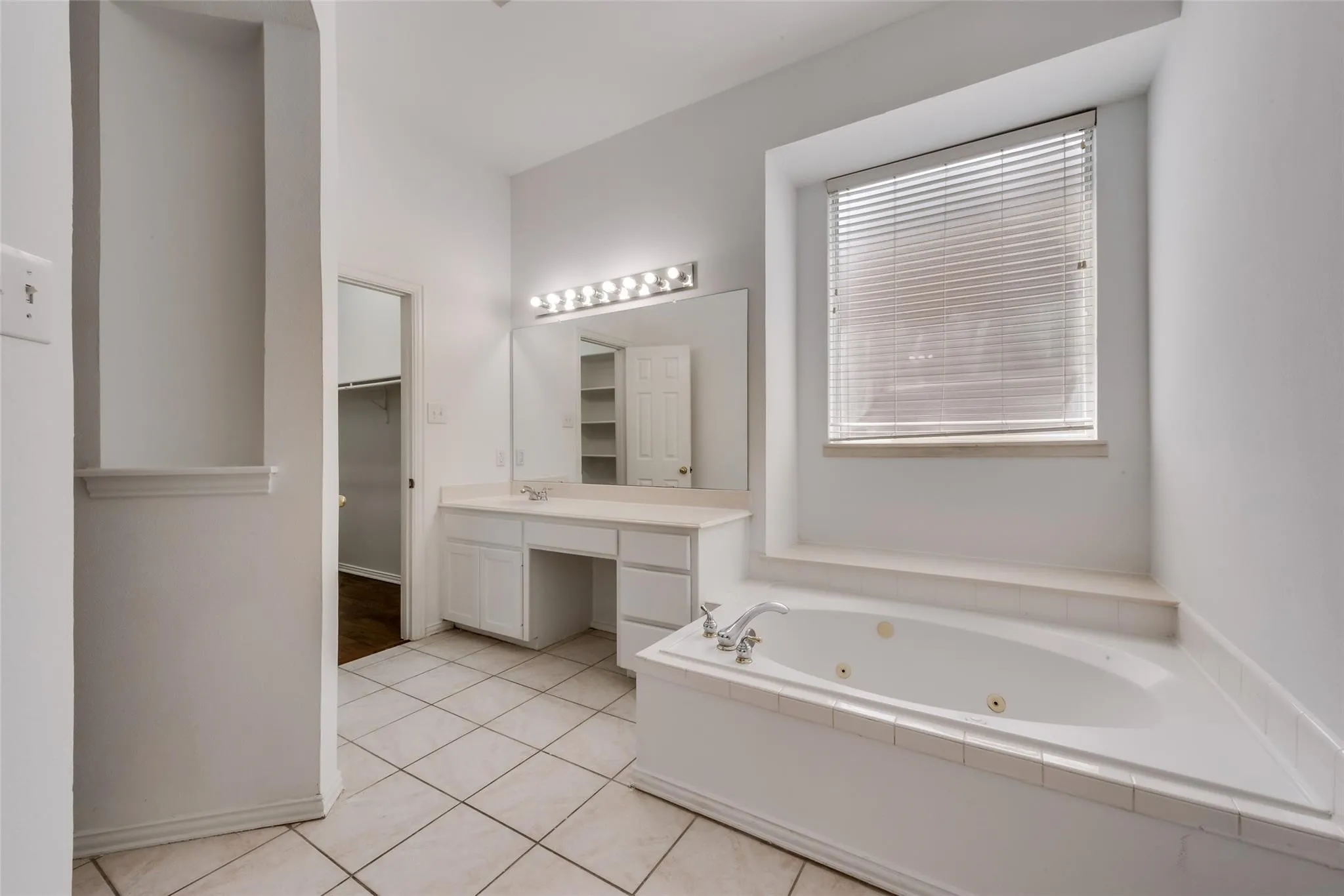 Bathroom featuring a built-in jetted tub with tile surround, white vanity with integrated sink, large mirror, and overhead vanity lighting