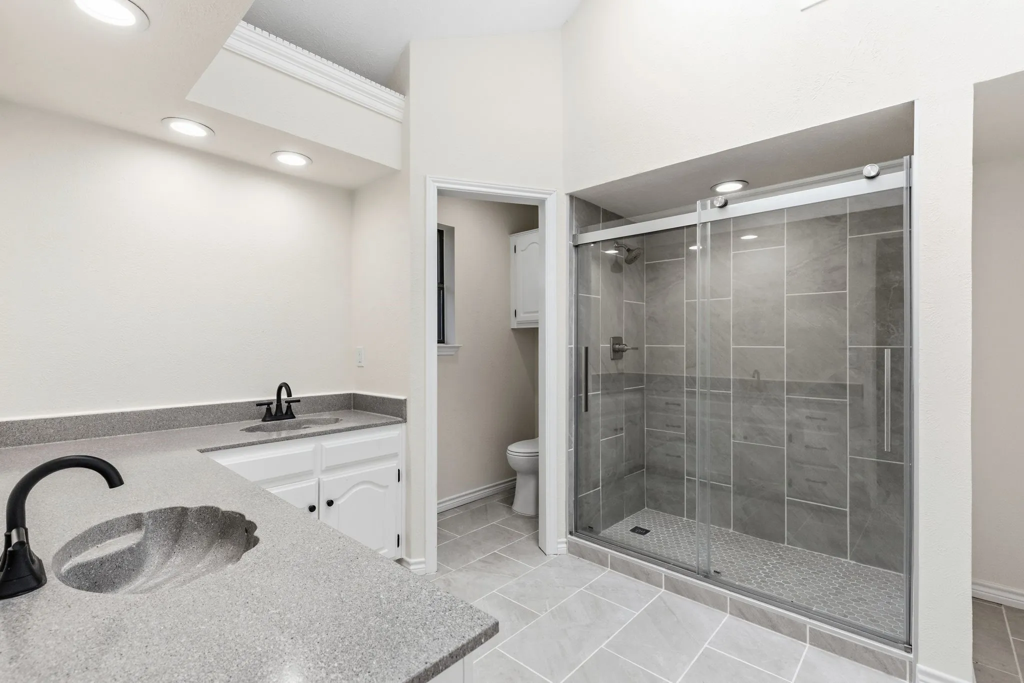 Bathroom featuring a glass-enclosed shower with large format tile surround, a built-in shower niche, dual vanities with light-colored countertops, a unique shell-shaped sink, and overhead recessed lighting
