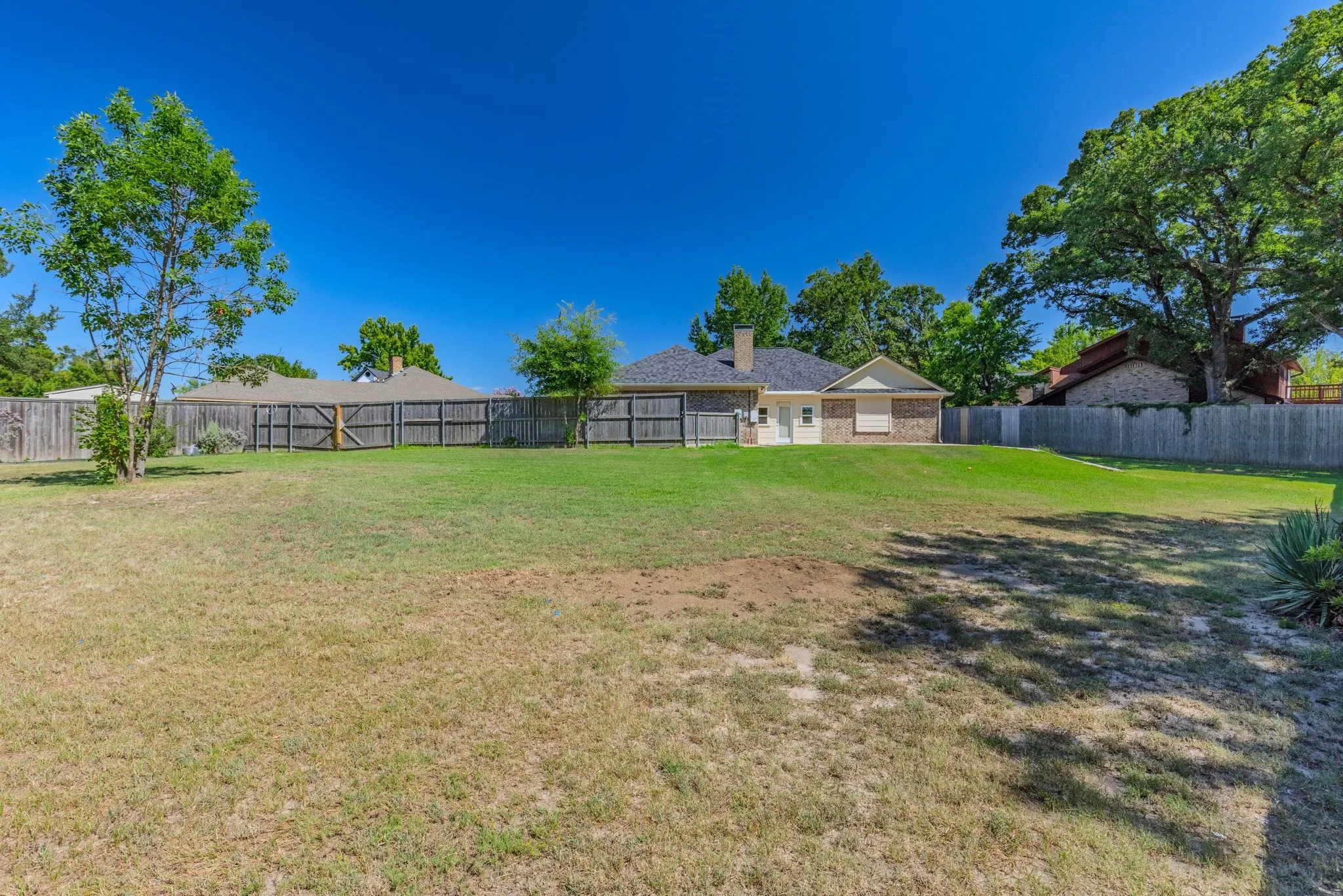 Expansive backyard featuring a partial wooden privacy fence, mature trees, and a brick-sided exterior with a white trim
