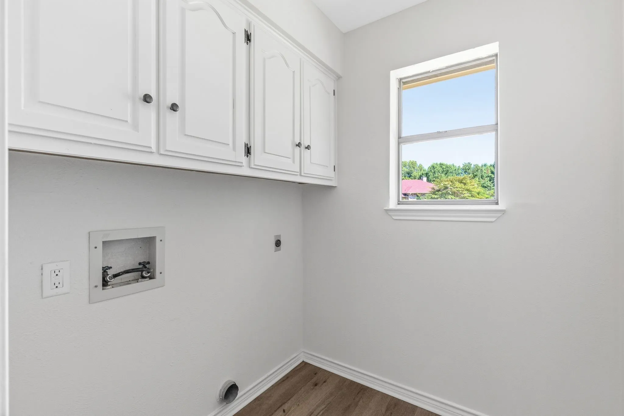 Dedicated utility space featuring overhead white cabinetry with black hardware, a single window, and wood-finish flooring