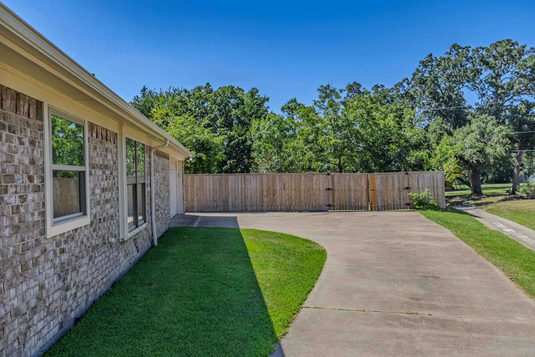 Brick exterior featuring multiple windows, a long concrete driveway, a wooden privacy fence, and a manicured lawn