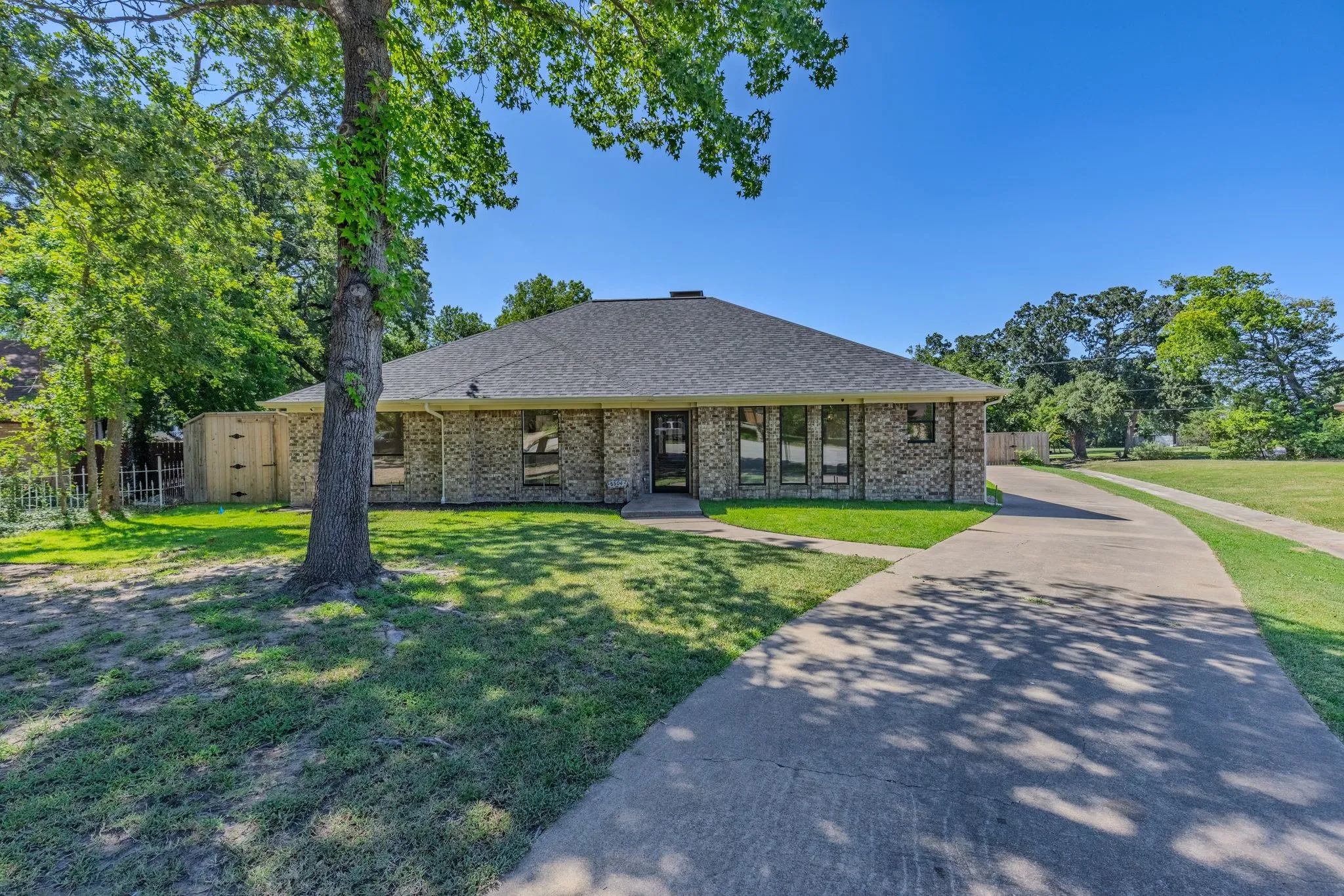 Brick exterior residential property with a gray shingle roof, featuring a concrete driveway, a large mature tree, and a green lawn