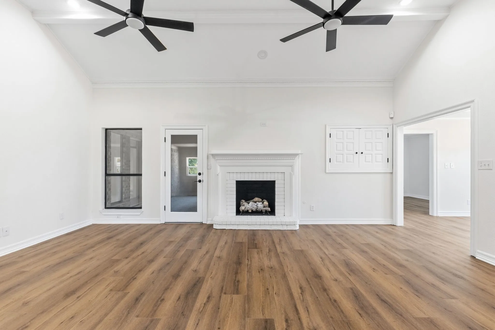 Spacious room featuring vaulted ceilings, wood-finish flooring, two modern ceiling fans, a white fireplace with a mantel, and a doorway opening to another interior space