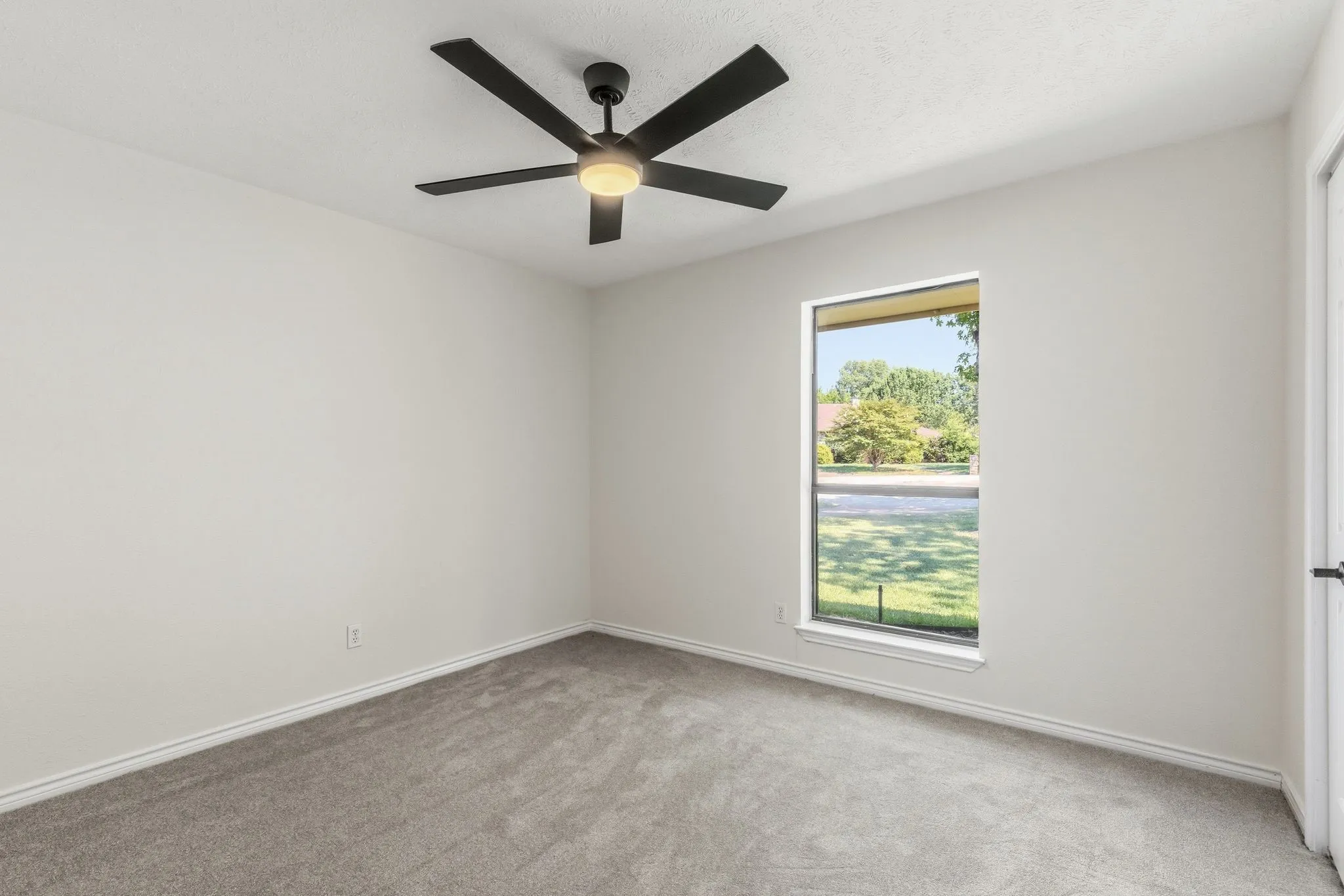 Neutral-toned room featuring a large window, gray carpeting, white baseboards, and a modern black ceiling fan with integrated lighting