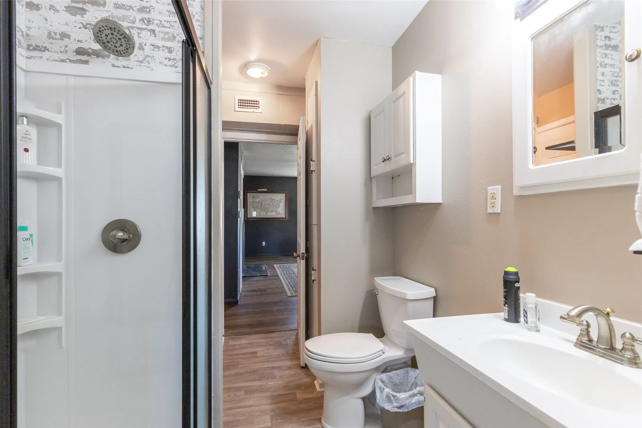 Bathroom featuring a shower with a brick-patterned surround, a white vanity with an integrated sink, a matching wall cabinet, and wood-finish flooring