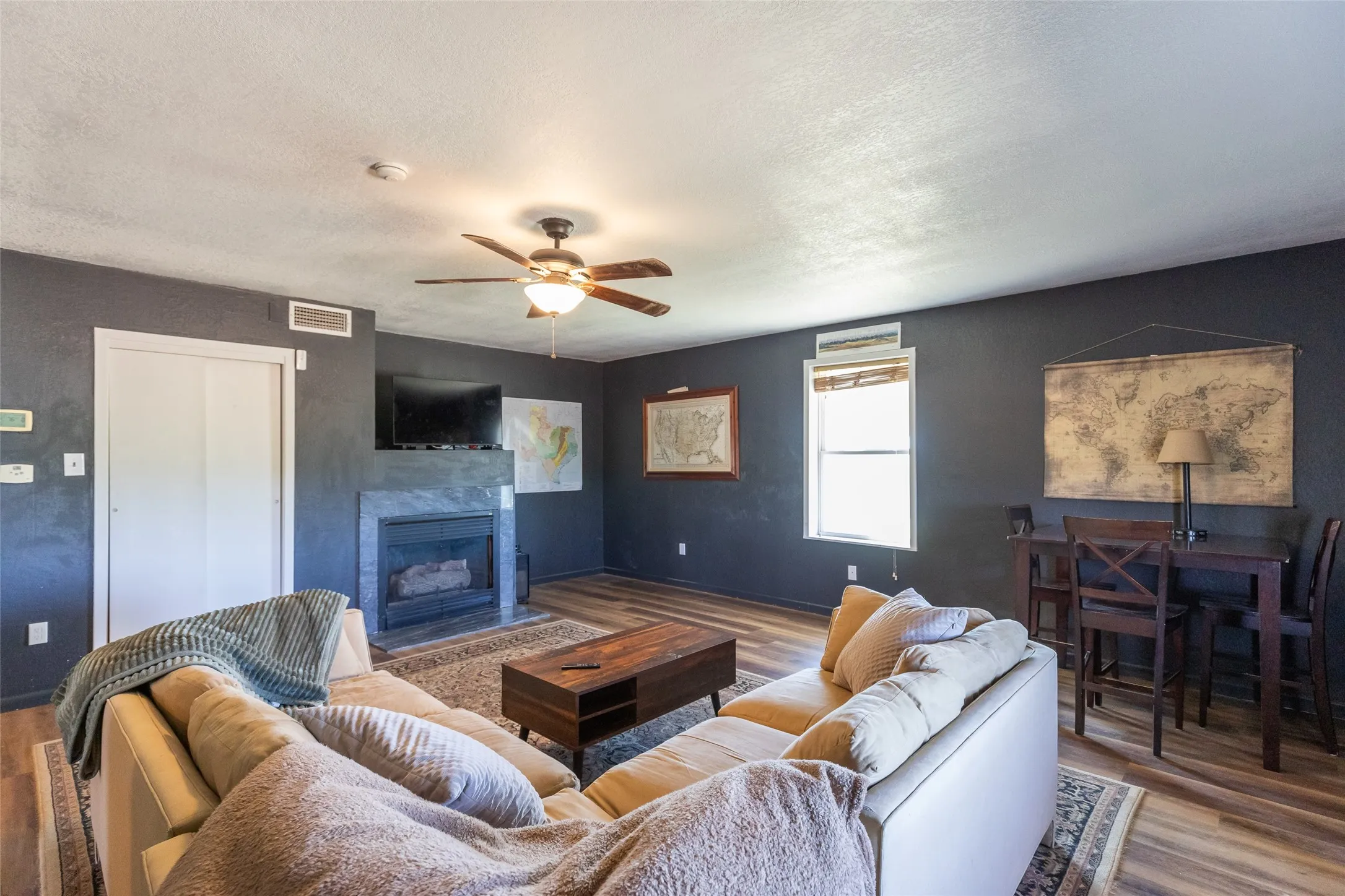Living area featuring wood-finish flooring, a ceiling fan, a fireplace with a stone-finish surround, and a window with blinds