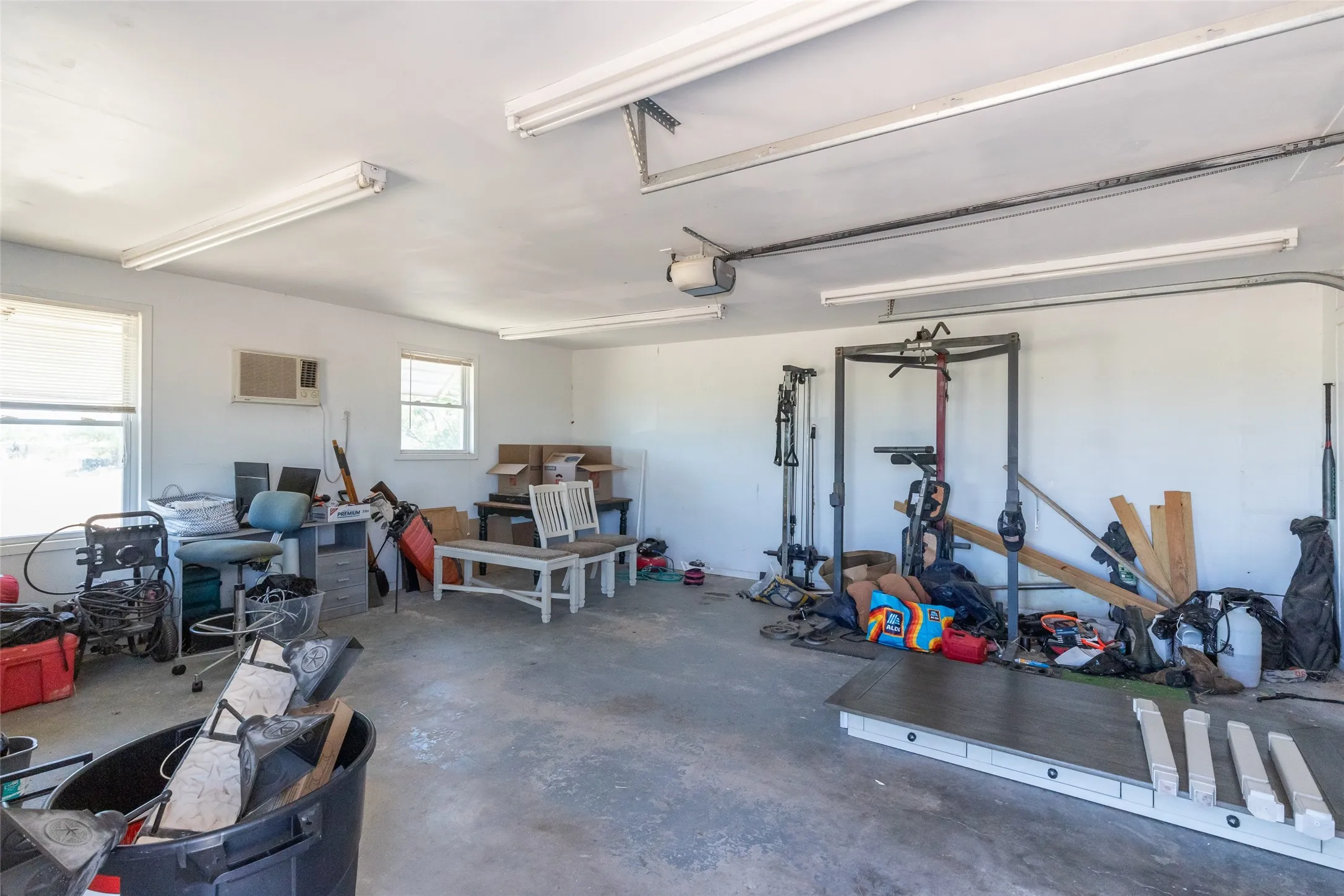 Spacious garage featuring a concrete floor, white painted walls, multiple windows for natural light, fluorescent overhead lighting, and a wall-mounted air conditioning unit