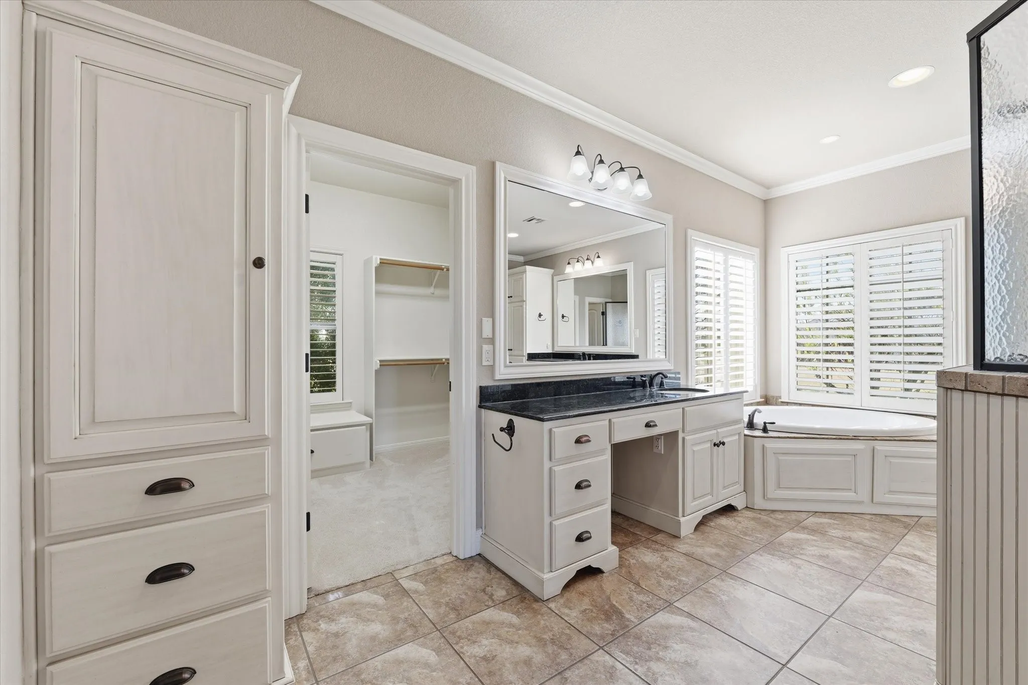 Spacious bathroom featuring tiled flooring, a built-in vanity with a dark countertop and ample storage, and a corner soaking tub with plantation shutters