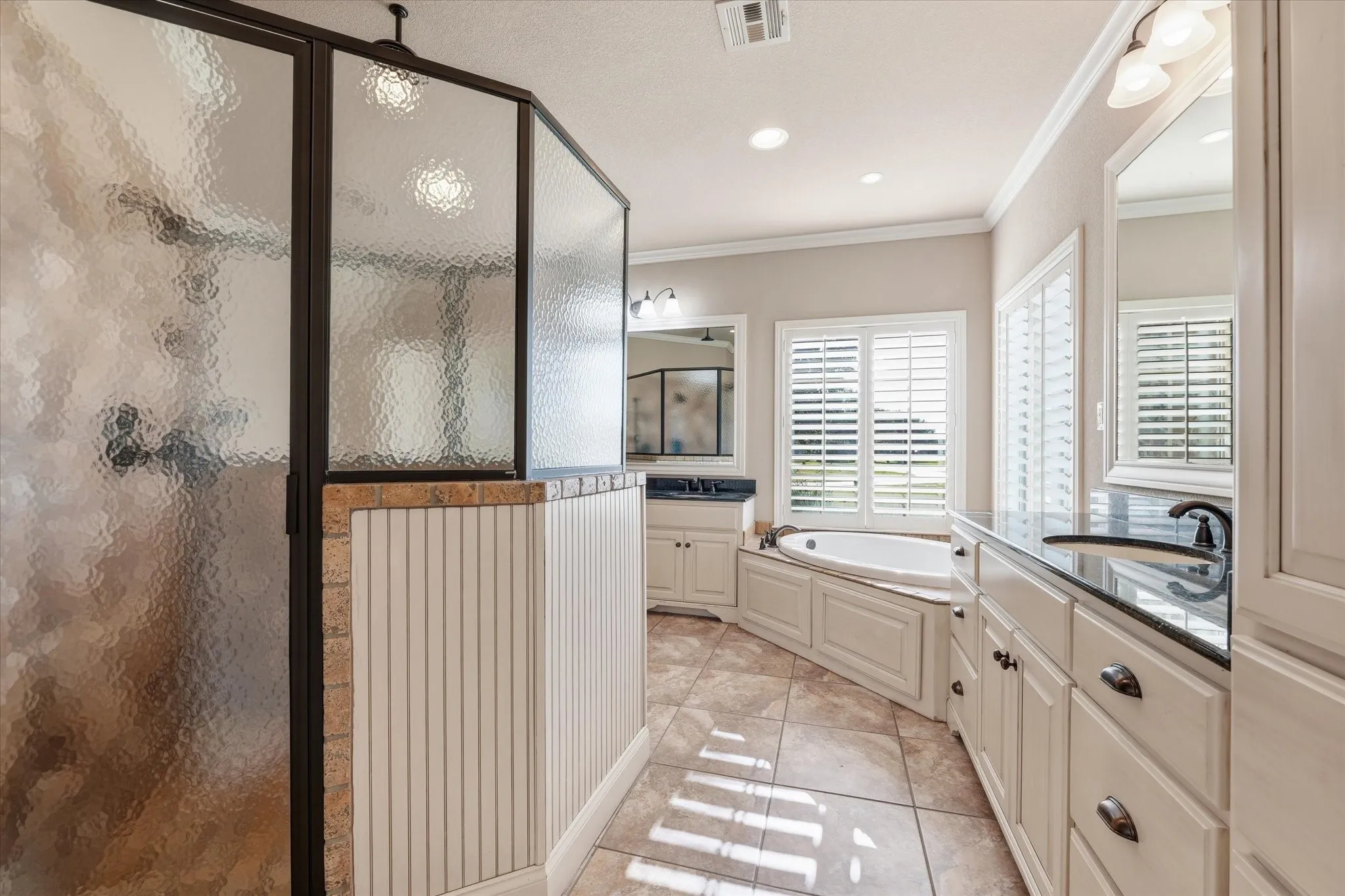 Enclosed shower with textured glass, corner soaking tub, dual vanity with black countertops, plantation shutters, and large format tile flooring