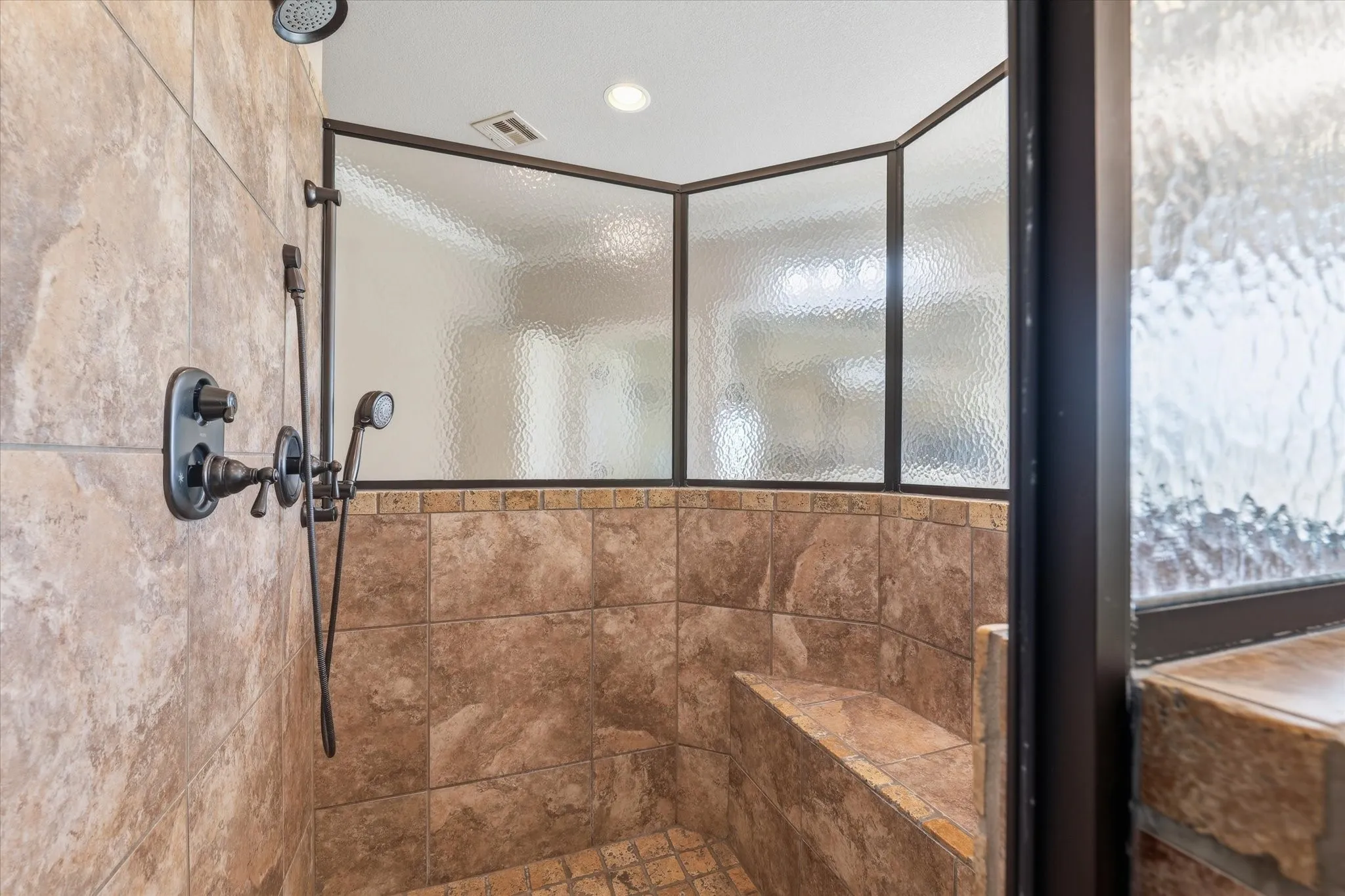 Walk-in shower featuring bronze fixtures, a built-in bench, textured privacy glass, ceiling-mounted shower head, and a handheld shower attachment