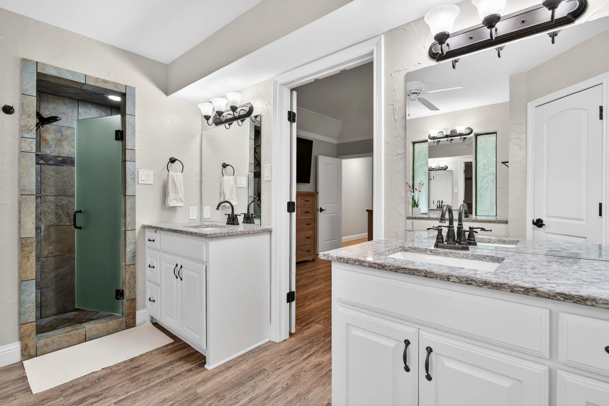 Bathroom featuring dual vanities with granite countertops and undermount sinks, a walk-in shower with stone-tiled walls and a frosted glass door, and wood-finish flooring