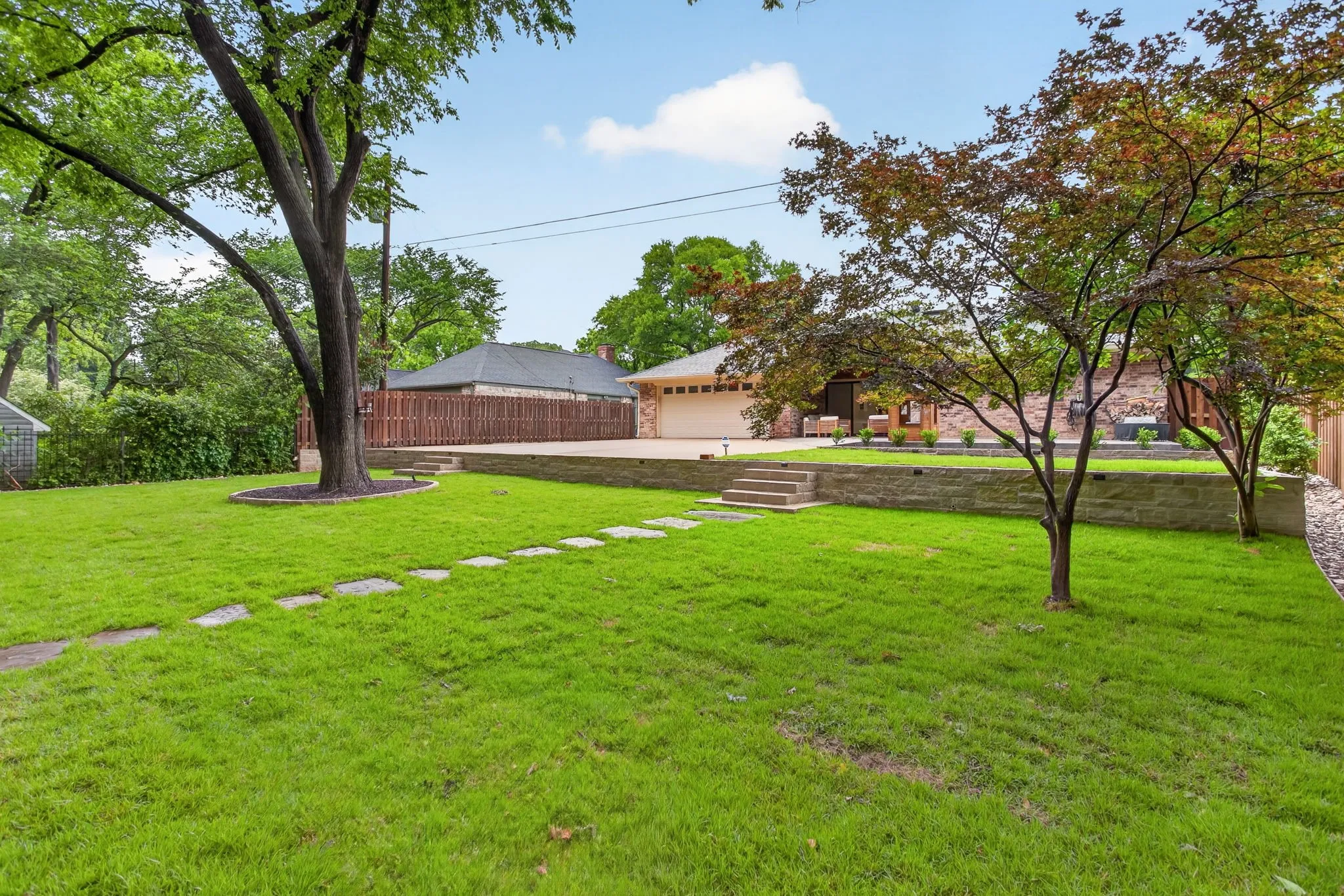 Expansive lawn featuring stepping stones, mature trees, a stone retaining wall, and a multi-level patio area