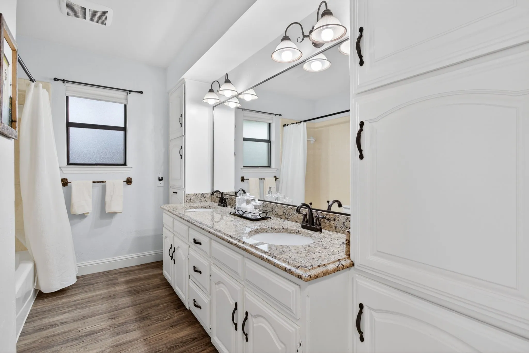 Guest Bathroom featuring a dual vanity with granite countertops, wood-finish flooring, white cabinetry with dark hardware, and a framed mirror with overhead lighting