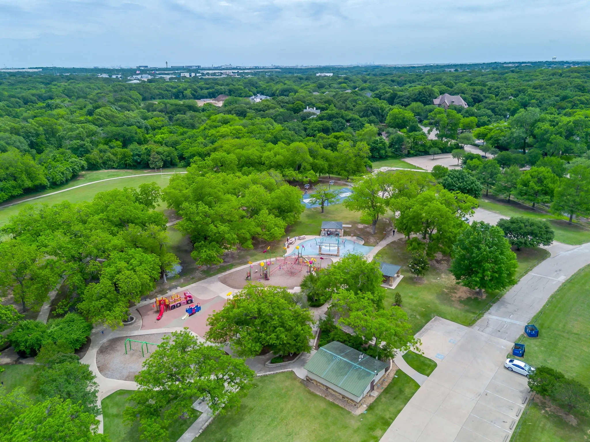 Splash pad, playground, and pavilions. Expansive park setting featuring mature trees, a splash pad, multiple play structures, pedestrian pathways, and a dedicated parking area