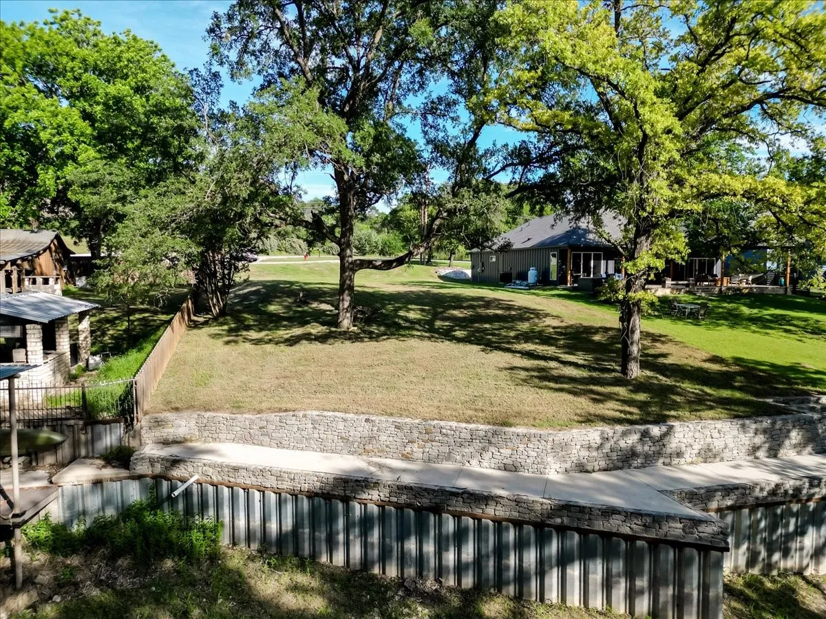 Expansive terraced yard with a stone retaining wall, mature trees, and a distant single-story dwelling featuring a dark roof and covered porch