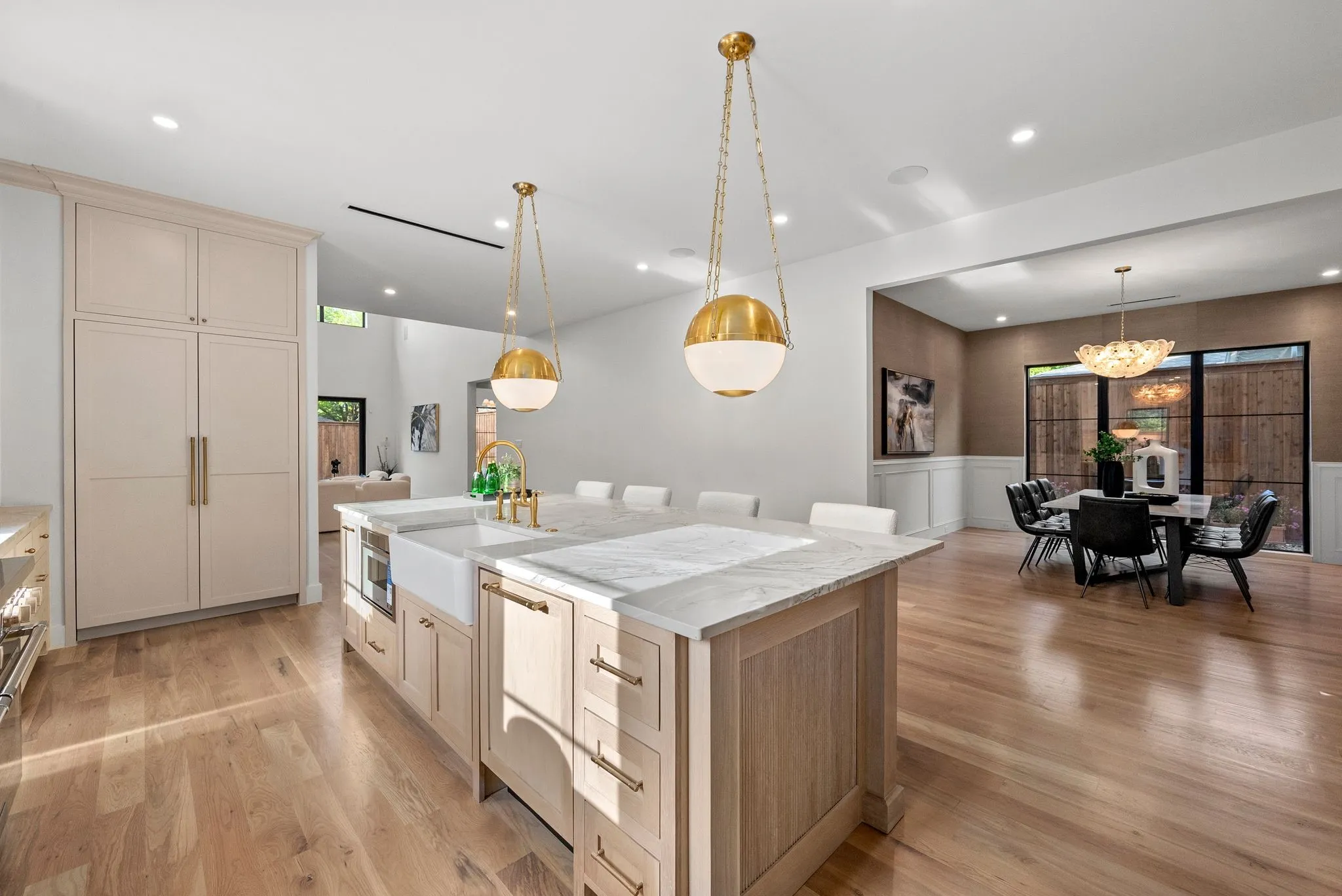 Gourmet kitchen featuring a large central island with a farmhouse sink, integrated appliances, gold-tone hardware, two spherical pendant lights, and wood-finish flooring