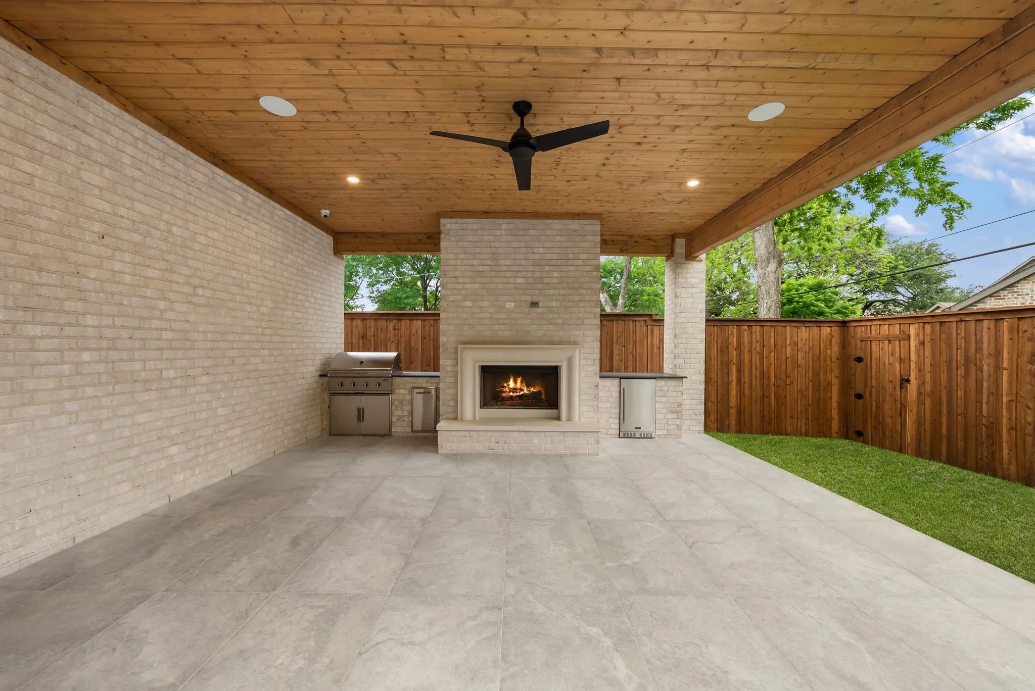 Covered outdoor patio featuring a brick-clad fireplace, built-in grill station, and wood-finish ceiling with recessed lighting