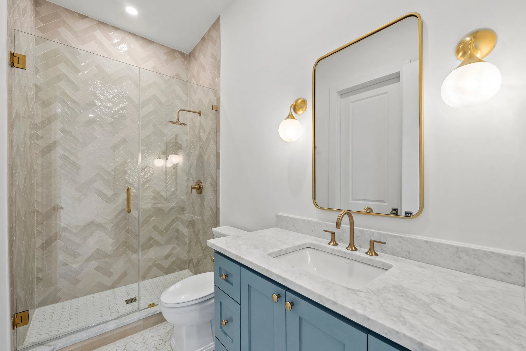Bathroom featuring a glass-enclosed shower with herringbone tile, a toilet, a light blue vanity with a white countertop, a rectangular mirror, and two wall-mounted sconces