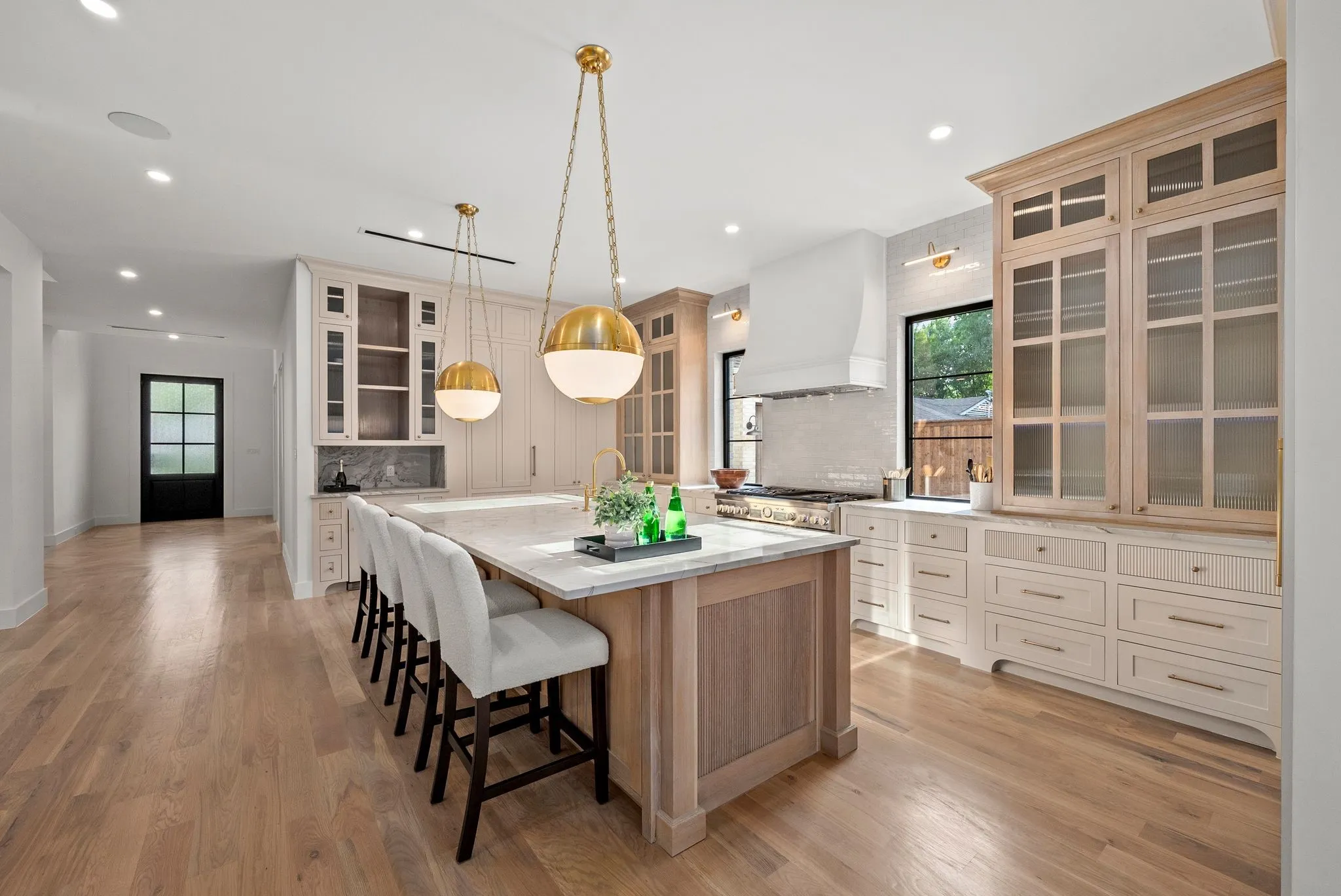 Spacious kitchen featuring a large central island with seating, brass pendant lighting, light wood-finish flooring, custom cabinetry with glass-front uppers, and stainless steel appliances