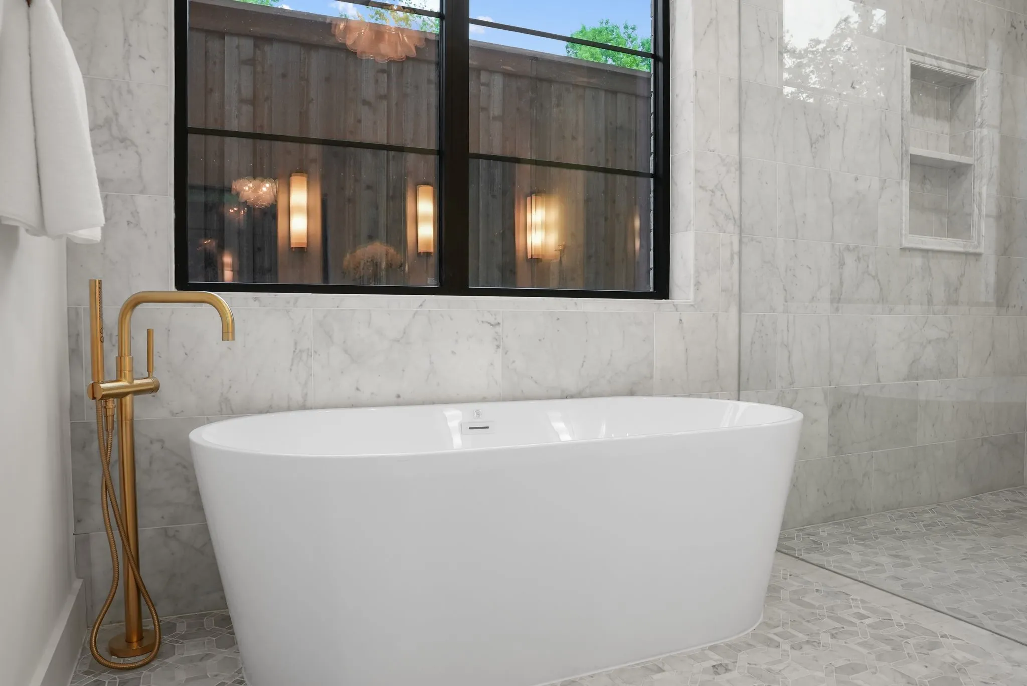 Freestanding soaking tub with a floor-mounted gold-tone faucet, set against large-format marble-look wall tiles and a pebble-style floor
