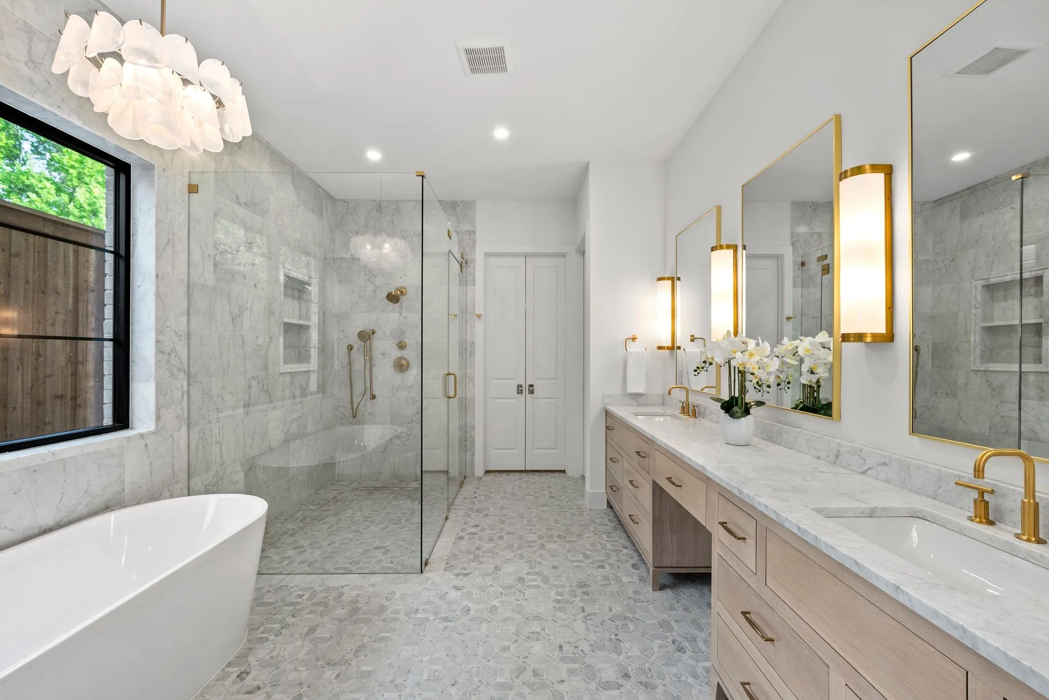 Spacious bathroom featuring a freestanding soaking tub, expansive glass-enclosed shower with bench and built-in niches, a dual vanity with light wood cabinetry, gold-framed mirrors, and pebble tile flooring