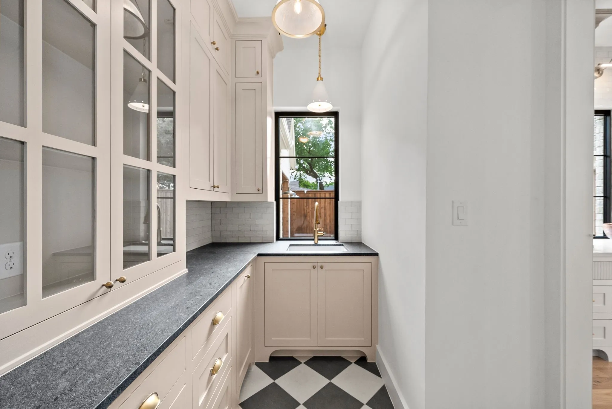 Built-in cabinetry with glass-front uppers and brass hardware, black and white checkered flooring, dark stone countertops, a subway tile backsplash, and a gold-finish faucet