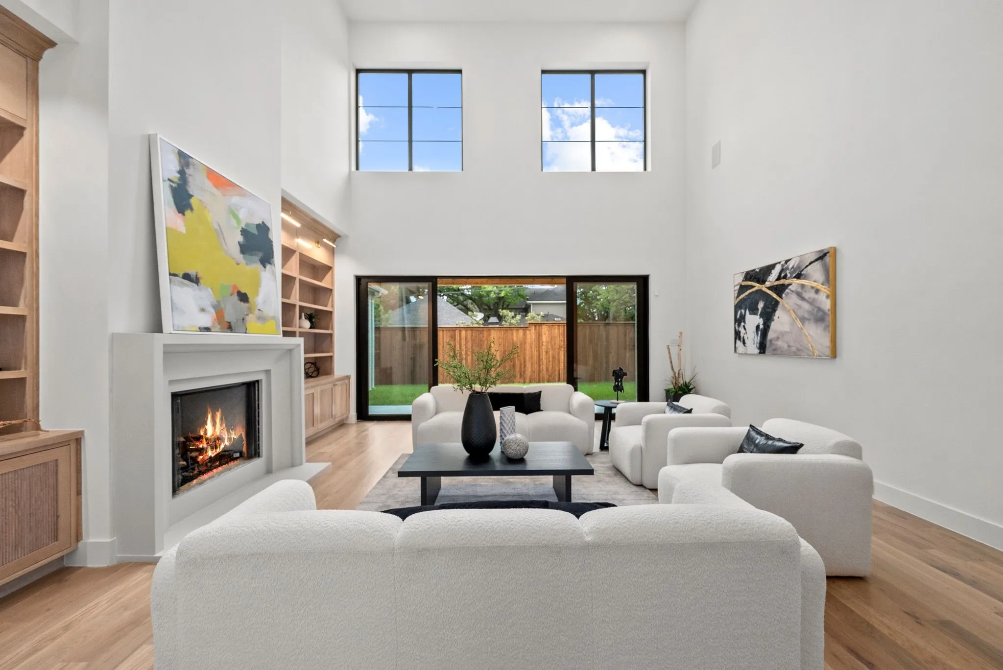 Spacious living area featuring a two-story ceiling, large fireplace with a white mantel, built-in natural wood shelving, expansive sliding glass doors, and light wood-finish flooring