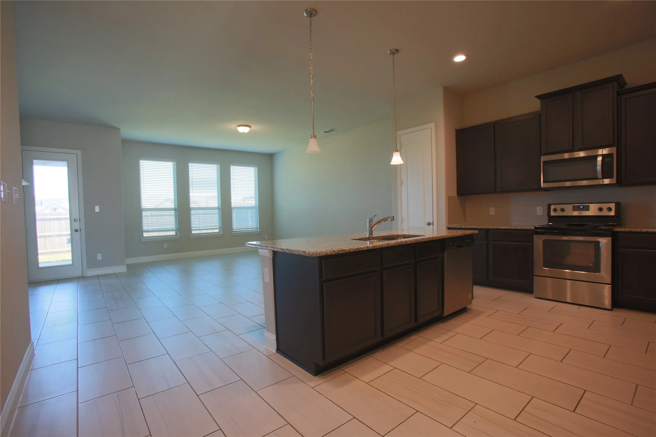 Open-concept kitchen and living area featuring plank-style tile flooring, a kitchen island with sink and pendant lighting, dark wood cabinetry, and stainless steel appliances